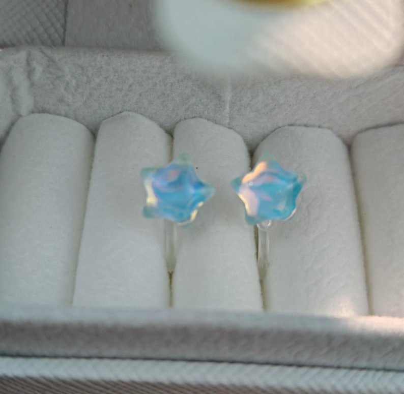 May include: A pair of star-shaped earrings with a light blue, iridescent finish. The earrings are displayed in a white jewelry box with a gray and white striped exterior. The earrings are small and delicate.