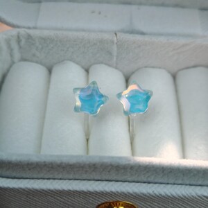 May include: A pair of star-shaped stud earrings. The earrings are made of clear, iridescent glass with a blue and white color scheme. They are displayed in a gray jewelry box with white padding.