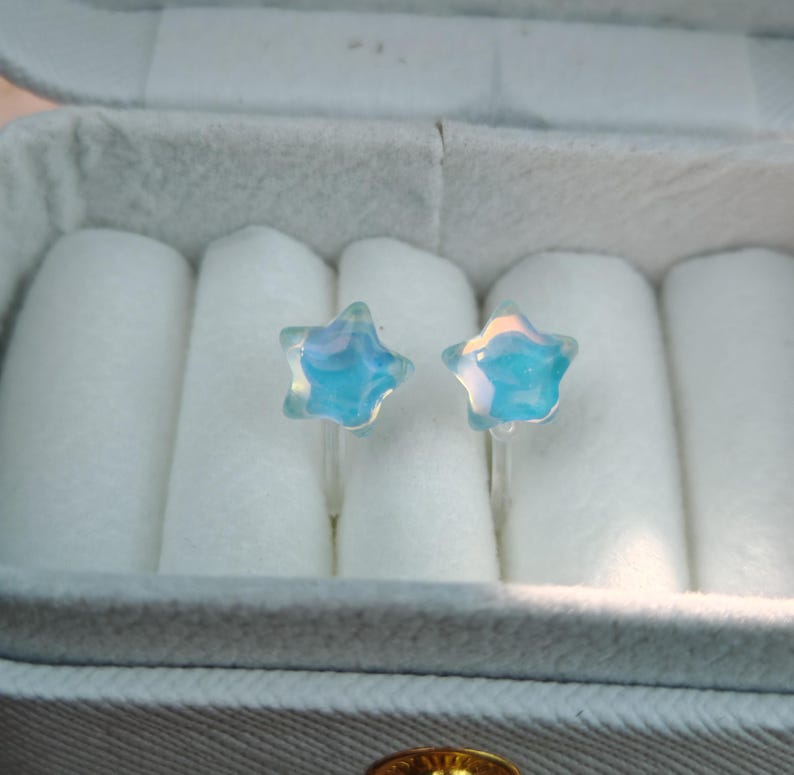 May include: A pair of star-shaped stud earrings. The earrings are made of clear, iridescent glass with a blue and white color scheme. They are displayed in a gray jewelry box with white padding.