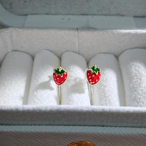 May include: A pair of strawberry-shaped earrings. Each earring features a red strawberry with green leaves and a gold-colored stem. The earrings are displayed in a white jewelry box with a light blue exterior.