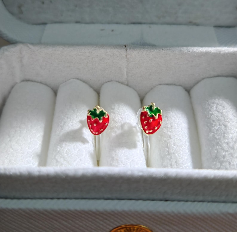 May include: A pair of strawberry-shaped earrings. Each earring features a red strawberry with green leaves and a gold-colored stem. The earrings are displayed in a white jewelry box with a light blue exterior.
