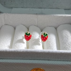 Puede incluir: Un par de pendientes de botón con forma de fresa. Los pendientes tienen un cuerpo rojo con motas blancas y una parte superior verde. Los pendientes se muestran en una caja de joyería blanca.