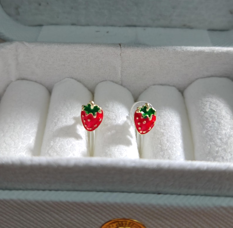 May include: A pair of strawberry-shaped stud earrings. Each earring features a red enamel strawberry with green leaves. The earrings are displayed in a white jewelry box with a light blue interior.