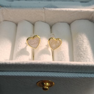 Puede incluir: Un par de pendientes de botón en forma de corazón. Los pendientes tienen un borde dorado y un diseño veteado en rosa claro y blanco. Se exhiben dentro de una caja de joyería blanca con exterior gris.