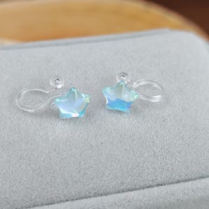 May include: A pair of iridescent star-shaped clip-on earrings. The earrings have a clear plastic clip and a light blue and white color scheme. The earrings are displayed on a gray surface.