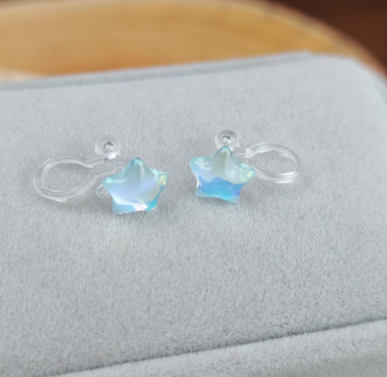 May include: A pair of iridescent star-shaped clip-on earrings. The earrings have a clear plastic clip and a light blue and white color scheme. The earrings are displayed on a gray surface.