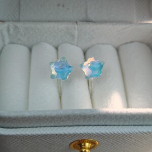 May include: A pair of star-shaped earrings with a light blue and iridescent finish. The earrings are displayed in a white jewelry box with a soft, velvety interior. The earrings are small and delicate.