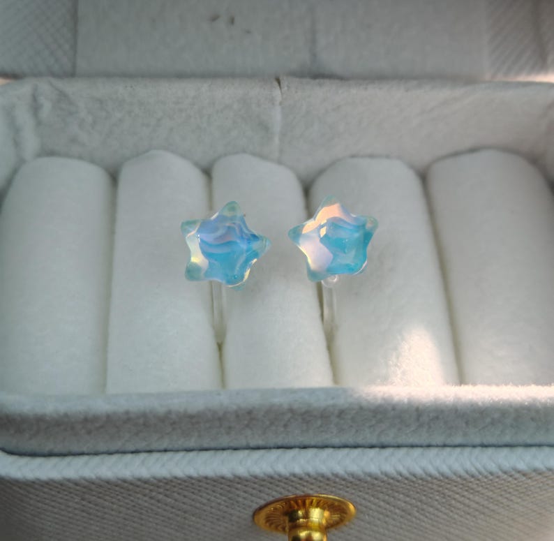 May include: A pair of star-shaped earrings with a light blue and iridescent finish. The earrings are displayed in a white jewelry box with a soft, velvety interior. The earrings are small and delicate.