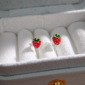 May include: A pair of strawberry-shaped stud earrings. Each earring features a red body with white seed details and a green top. The earrings are displayed in a white jewelry box.