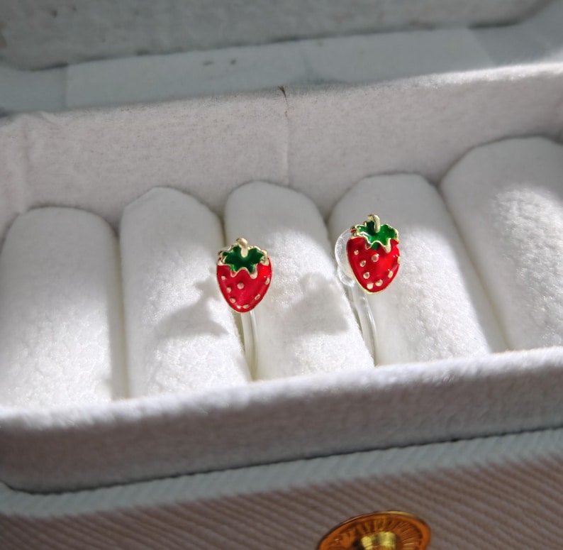 May include: A pair of strawberry-shaped stud earrings. Each earring features a red body with white seed details and a green top. The earrings are displayed in a white jewelry box.