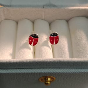 Puede incluir: Un par de pendientes de botón con forma de mariquita. Cada pendiente tiene un cuerpo rojo con lunares negros y una cabeza negra. Los pendientes se muestran en una caja de joyería gris con un interior blanco acolchado.