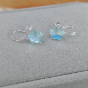 May include: A pair of clip-on earrings featuring iridescent star-shaped charms. The stars have a light blue and white color scheme, with a translucent appearance. The clip-on mechanisms are clear plastic.