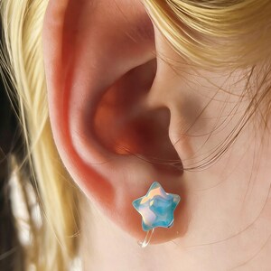 May include: A close-up of a person's ear with a star-shaped earring. The earring is a light blue color with iridescent highlights. The earring is attached to the ear with a clear, thin wire.