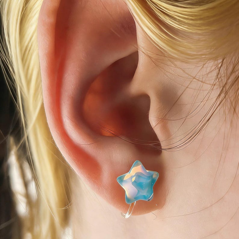 May include: A close-up of a person's ear with a star-shaped earring. The earring is a light blue color with iridescent highlights. The earring is attached to the ear with a clear, thin wire.