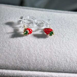 May include: A pair of clip-on earrings featuring a red strawberry design with green leaves and gold accents. The earrings are attached to clear plastic clips, resting on a gray surface.
