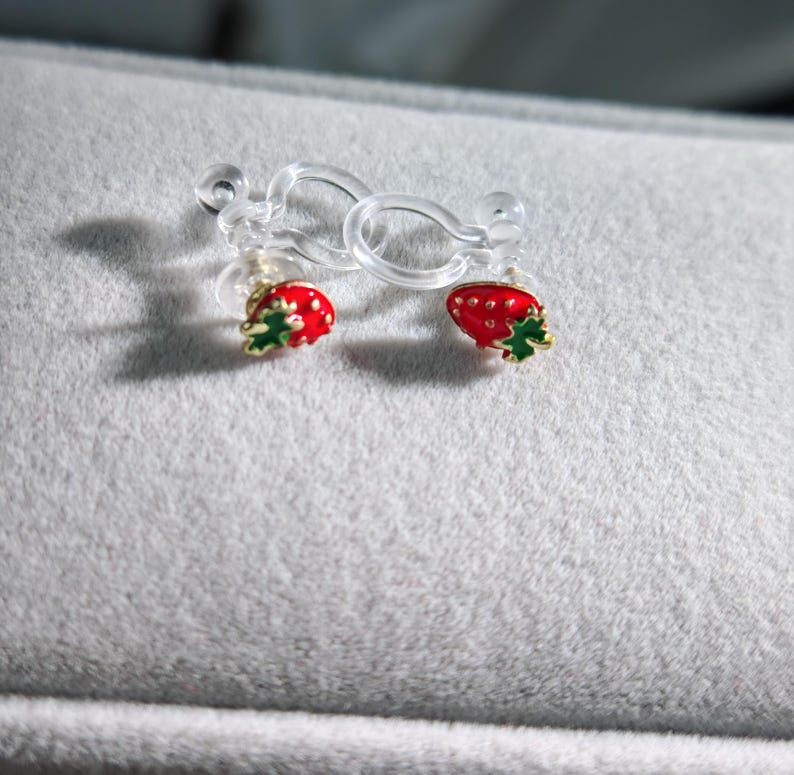 May include: A pair of clip-on earrings featuring a red strawberry design with green leaves and gold accents. The earrings are attached to clear plastic clips, resting on a gray surface.