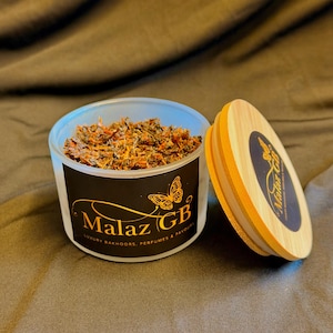 May include: A clear container filled with dried herbs and spices, with a wooden lid beside it. The container has a black label with gold text and a butterfly graphic. The label reads "Malaz GB Luxury Bakhoors, Perfumes & Favours".