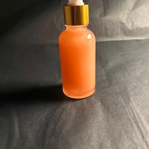 May include: A clear glass dropper bottle containing a peach-coloured liquid. The bottle has a gold-coloured collar and a white dropper. The background is a dark grey colour.