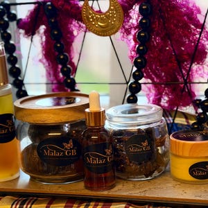 May include: A selection of beauty products in glass jars and bottles with gold and wooden details. The products are labelled with "Malaz GB" and a butterfly logo. The background includes decorative elements, with a focus on the products.