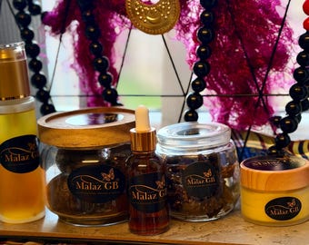 May include: A selection of beauty products in glass jars and bottles with gold and wooden details. The products are labelled with "Malaz GB" and a butterfly logo. The background includes decorative elements, with a focus on the products.