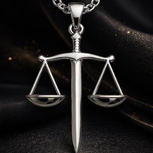 May include: A silver-colored pendant necklace featuring a stylized sword with a scale design. The pendant hangs from a matching chain. The scales of justice are balanced on either side of the sword.