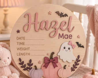 May include: A round wooden birth announcement sign featuring the name Hazel Mae in pink script. The sign includes engraved lines for date, time, weight, and length, decorated with a white ghost, pink pumpkins, bats, and stars.