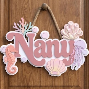 May include: A wooden door sign with the name "Nany" in pink, surrounded by sea-themed decorations. Includes a seahorse, jellyfish, coral, and seashells. The sign is suspended by a rope and a wooden knob.