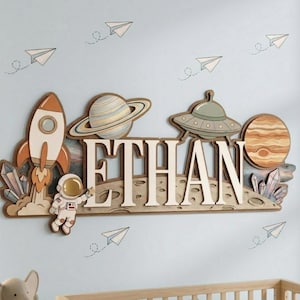 Könnte beinhalten: Eine Holz-Wanddekoration mit dem Namen "ETHAN" in großen, hellen Buchstaben, mit Weltraum-Thema. Das Design umfasst eine Rakete, Planeten, einen Astronauten und ein UFO, alles vor einem hellblauen Hintergrund mit Papierflieger-Akzenten.
