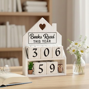 May include: A white wooden block calendar with the text "Books Read This Year." The calendar displays the numbers 306 and 59. A small potted plant and an owl figurine are on the shelf. A vase of daisies and an open book with glasses are also present.
