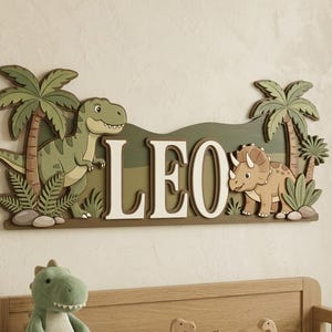 May include: A wooden sign with the name "LEO" in white letters, featuring cartoon dinosaurs and palm trees. The sign is in shades of green and brown, with a T-Rex on the left and a Triceratops on the right. A plush dinosaur and wooden toys are below.