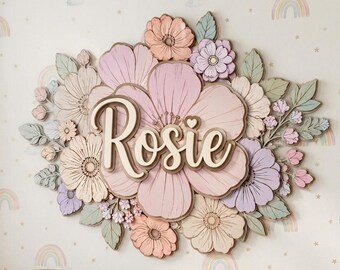 May include: A layered wooden wall sign featuring the name Rosie in cursive script, centered on a large pink flower. The design is surrounded by various smaller flowers and leaves in shades of peach, lavender, cream, and sage green.
