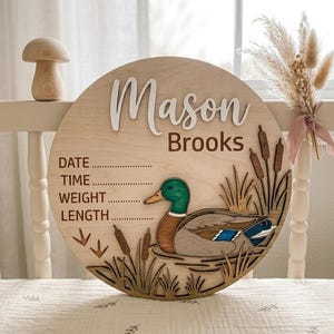 以下が含まれることがあります： Mason Brooksという名前が刻まれた円形の木製出生記録ボード。日付、時間、体重、身長を記入する線が印刷されています。ボードには、池に浮かぶマガモとガマの穂、草がレイヤー状のイラストで描かれています。