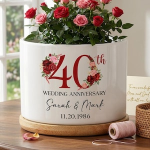 May include: A white ceramic planter with a floral arrangement of red and pink roses. The planter is decorated with the text "40th WEDDING ANNIVERSARY Sarah & Mark 11.20.1986". The planter sits on a wooden base.