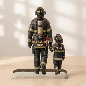 May include: A wooden figurine depicting a firefighter and a child in matching uniforms, walking together. The adult figure has "FIREFIGHTER MARK" on the back. The child's uniform reads "ETHAN". The figures are in black, gold, and brown tones.