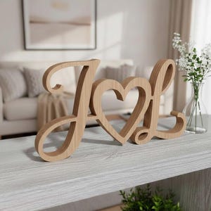 Puede incluir: Letrero de madera "Love" con la letra "O" reemplazada por un corazón. El letrero está hecho de madera de color claro y se encuentra sobre una superficie gris. Un jarrón con flores blancas está en el fondo.