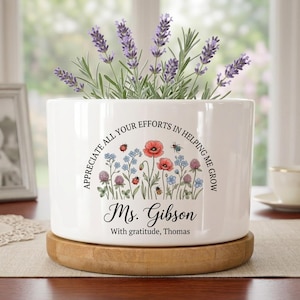 Puede incluir: Una maceta de cerámica blanca con base de madera, llena de lavanda y con un diseño floral y el texto "Appreciate all your efforts in helping me grow" y "Ms. Gibson".