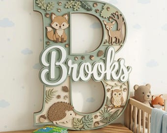 May include: A wooden letter "B" with the name "Brooks" in white script, decorated with woodland creatures like a fox, deer, owl, and hedgehog. The letter is painted in shades of green and natural wood tones, with leaf and acorn accents.