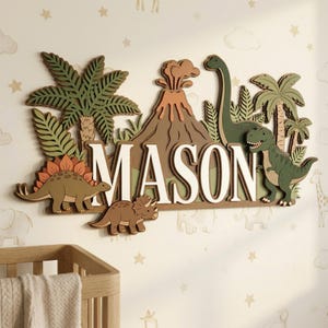 May include: Wooden wall decoration spelling "MASON" in white, with dinosaur and prehistoric landscape cutouts. Features a volcano, palm trees, and dinosaurs in green, brown, and orange. The decor is designed for a child's room.