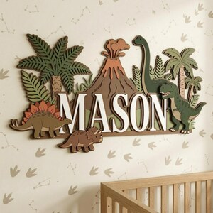 May include: A wooden wall decoration spelling "MASON" in white, with a dinosaur theme. The design includes a volcano, palm trees, and various dinosaur figures in shades of brown, green, and orange. The background is a light-coloured wall with a constellation pattern.