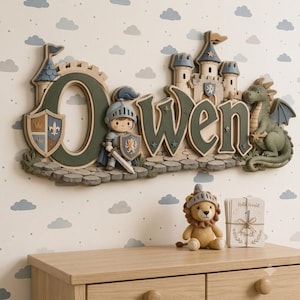 May include: A wall-mounted wooden name sign spelling Owen in dark green letters, featuring a knight with a sword and shield, a stone castle, and a green dragon. Below the sign sits a small knitted lion plush and a card labeled Hello World 1 Month.