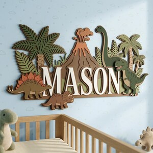 May include: A wooden wall decoration spelling "MASON" in white, with a dinosaur theme. It features a volcano, palm trees, and dinosaurs in green, brown, and orange. The background is light blue with cloud and star details, suitable for a child's room.