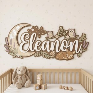 May include: A wooden nursery name sign featuring the name "Eleanor" in white script. The sign includes a crescent moon, stars, owls, a sleeping fawn, and floral accents in shades of brown, white, and lavender.