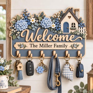 May include: A wooden wall-mounted key holder featuring a house illustration, blue hydrangea and white daisy floral accents, and the text "Welcome" and "The Miller Family". It includes six metal hooks holding various keys, a leash, and accessories.