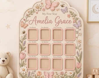 Puede incluir: Un tablero de fotos de madera clara titulado My First Year con el nombre Amelia Grace. Cuenta con doce marcos rectangulares para fotos mensuales, decorados con mariposas, abejas y enredaderas florales en relieve en tonos rosa y verde.