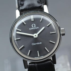 Pode incluir: Um close-up de um relógio Omega Genève vintage com mostrador preto e ponteiros e marcadores prateados. O relógio tem uma caixa prateada e uma pulseira de couro preta. O mostrador exibe o logotipo da Omega e a palavra "Genève".