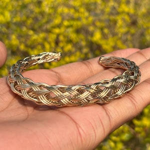 May include: A gold-coloured woven cuff bracelet. The bracelet is held in an open hand against a backdrop of green and yellow foliage. The cuff is made of interwoven strands, creating a textured, open design. The bracelet is a statement piece.