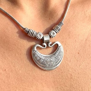May include: A silver necklace with a crescent pendant. The pendant has an intricate geometric design and hangs from a delicate chain. Decorative elements are near the clasp. The necklace is a piece of jewellery.