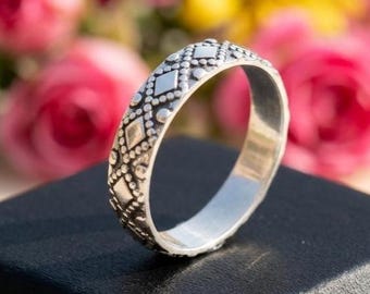 May include: A silver ring with a detailed pattern of diamonds and dots. The ring is displayed on a black surface with a blurred background of pink and yellow flowers. The ring's design is intricate and eye-catching, suitable for everyday wear or special occasions.