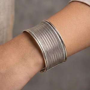 Op de afbeelding: Een brede zilveren manchetarmband met ingewikkelde, getextureerde details. De armband heeft een reeks parallelle lijnen en een decoratieve rand. De sieraden worden om een arm gedragen en tonen het ontwerp en vakmanschap.