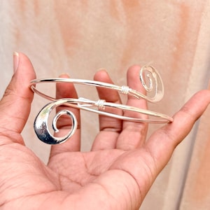 May include: A silver-toned metal cuff bracelet featuring a bypass design with two open ends. Each end of the bracelet terminates in a flat, polished spiral shape. The band is constructed from a smooth, rounded wire that wraps around the wrist.